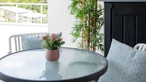 A round glass table with a pink pot of flowers, white chairs with patterned cushions, and a bamboo plant beside a black wall.