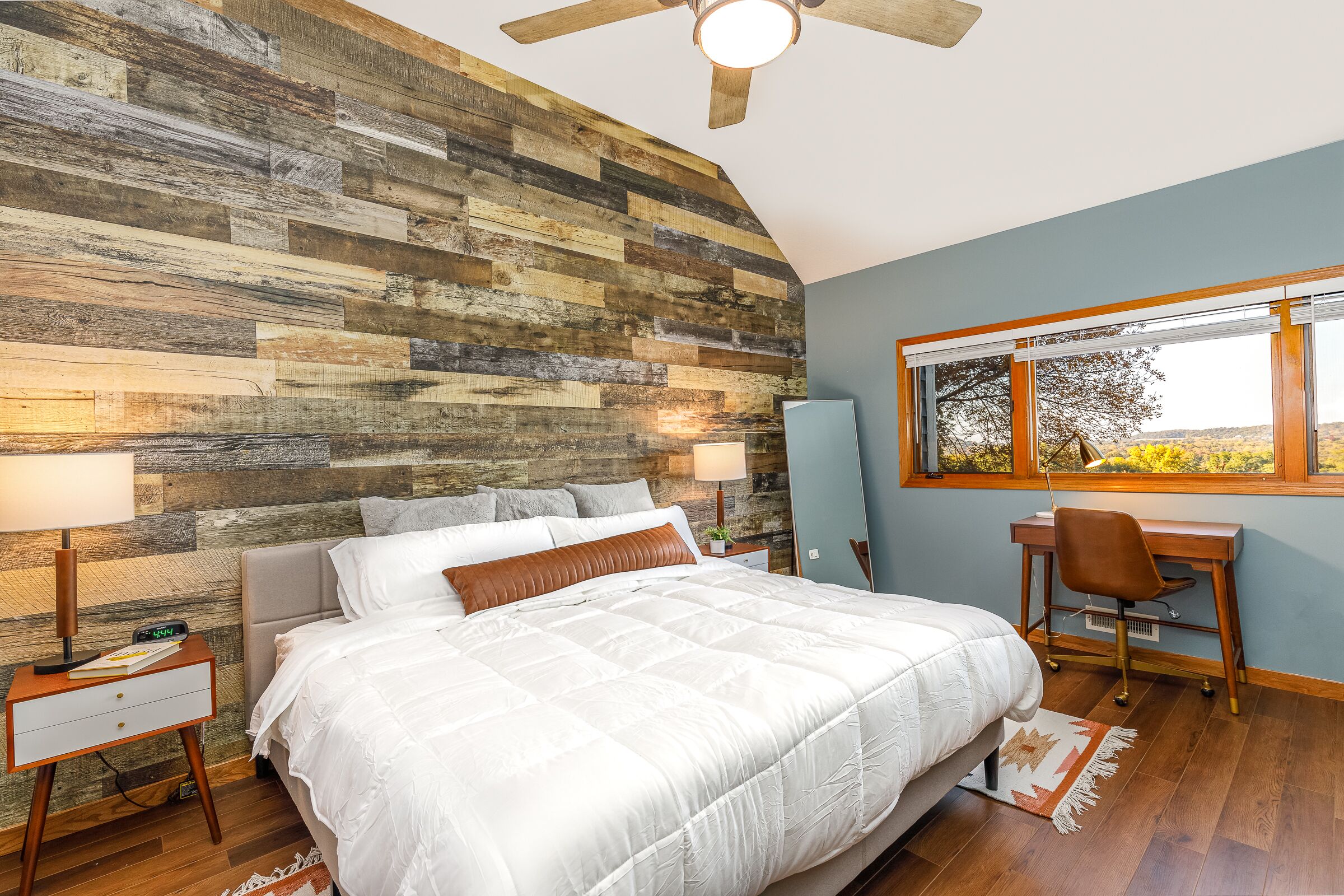 A bedroom with a wooden wall, a bed with white bedding, a desk with a chair, and a window with a view.