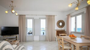 A modern living room with a flat-screen TV, a dining table with chairs, a fireplace, and a view of the sea through large windows. - Christiana Hotel (Paros)