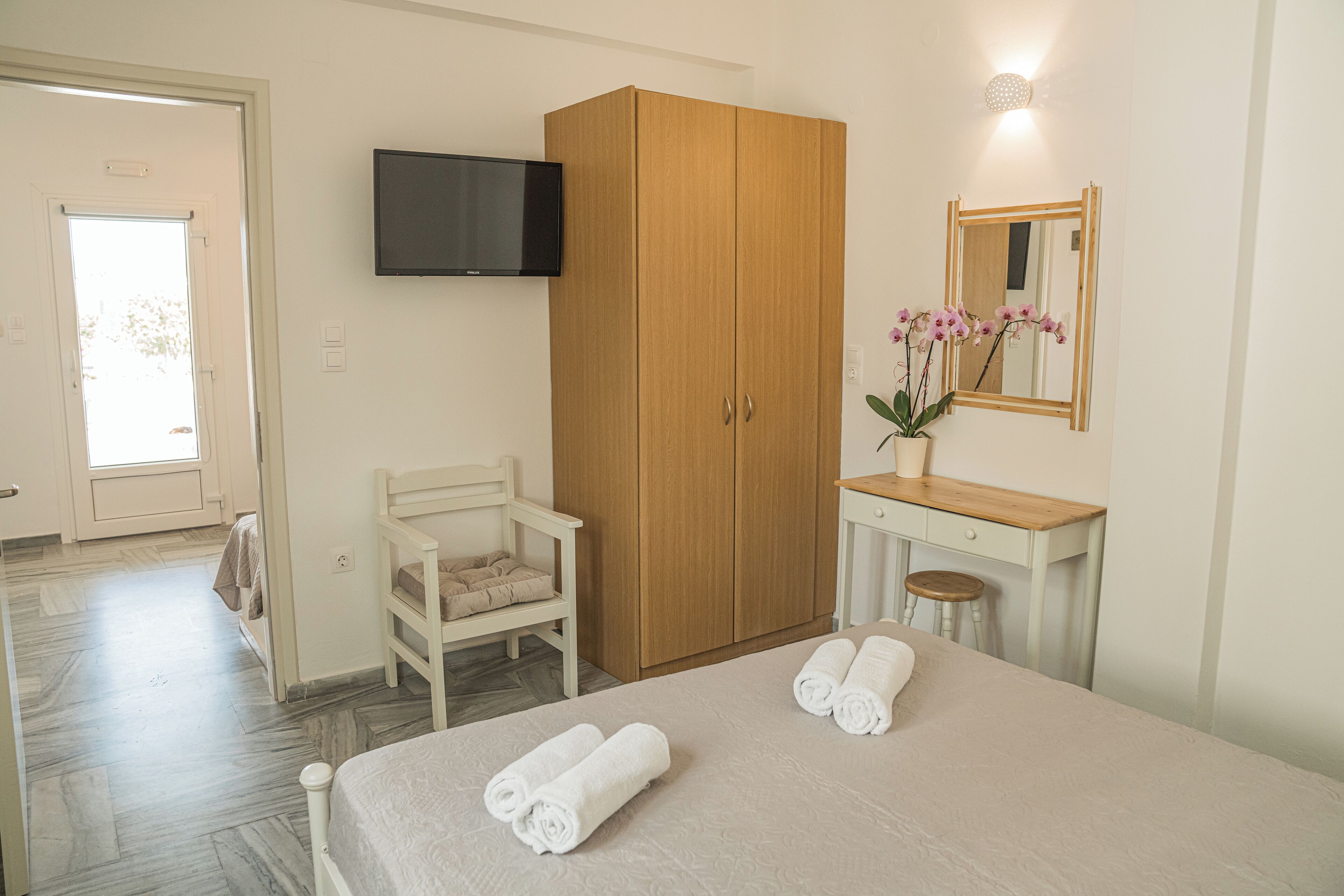 A bedroom with a bed, a wooden wardrobe, a television, a desk with a mirror, and a chair.