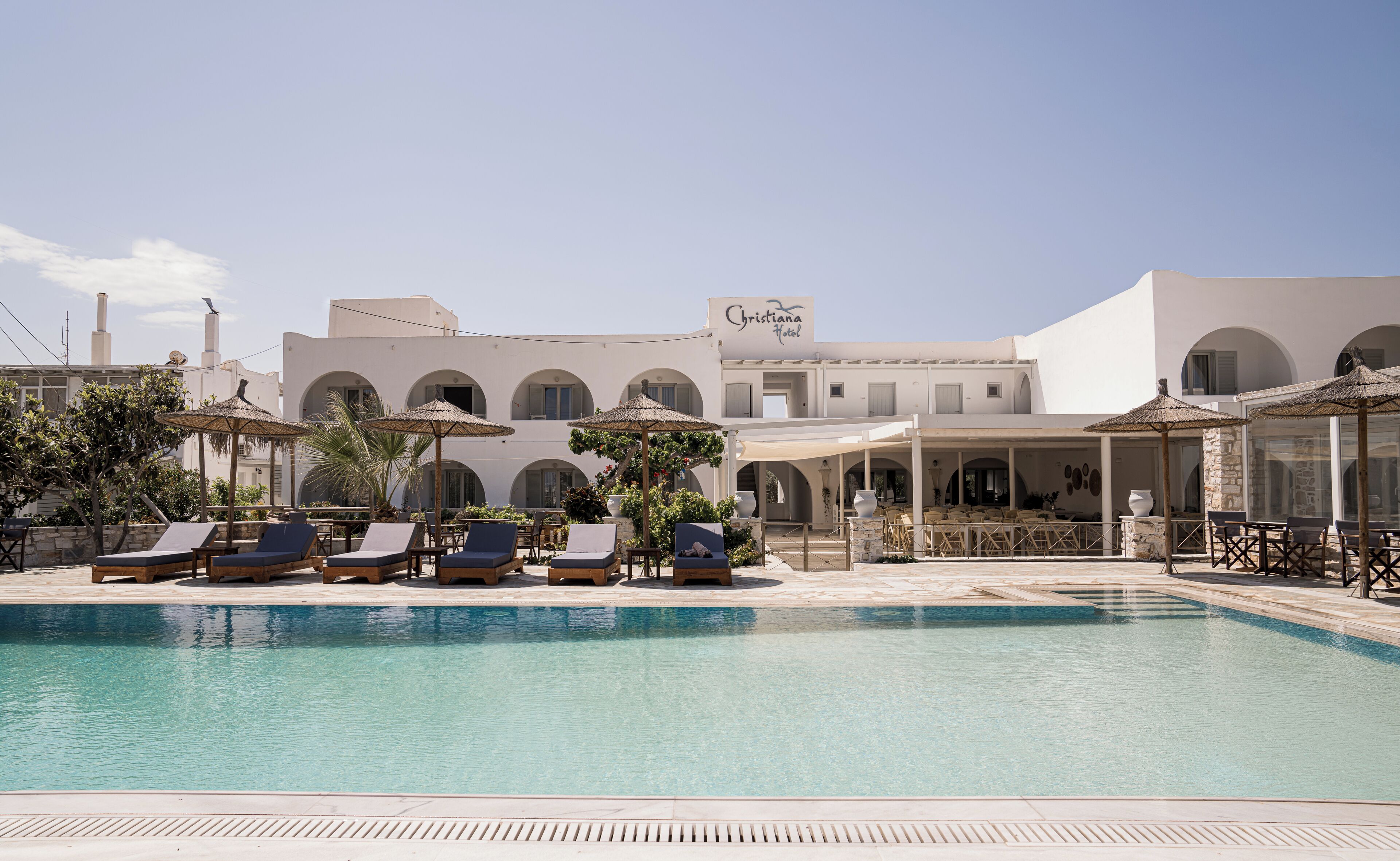 A pool area with lounge chairs and umbrellas, a sign for 'Christiana Hotel', and a building with arches and a white facade.