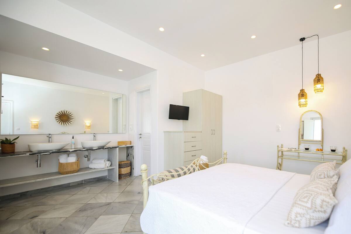 A modern bedroom with a large bed, a vanity with two sinks, a wardrobe, and a television mounted on the wall.
