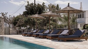 A poolside area with lounge chairs and umbrellas, surrounded by trees and a stone building. - Christiana Hotel (Paros)
