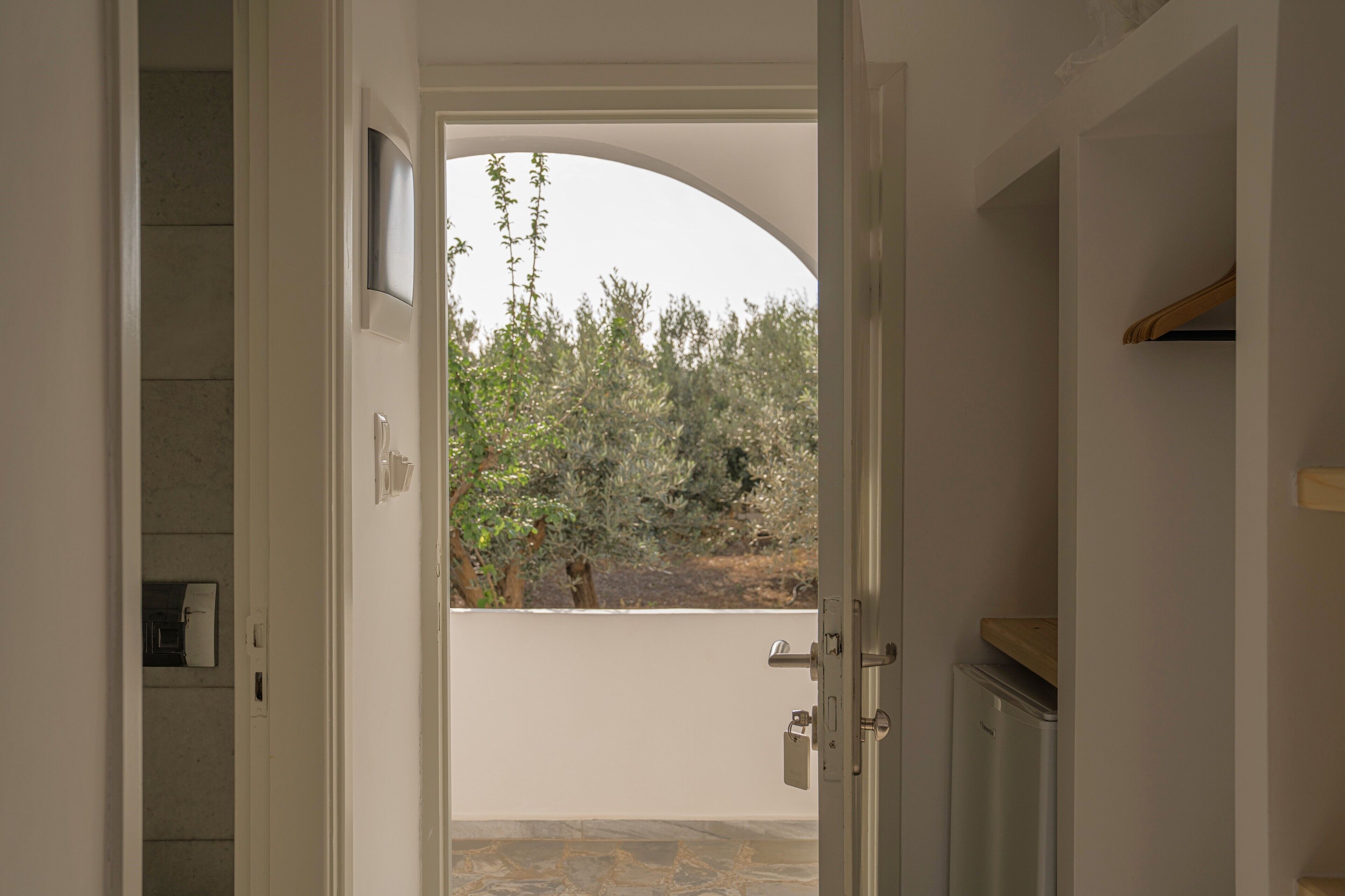 Une porte ouverte donnant sur un balcon d’où l’on peut admirer des arbres et un mur blanc.