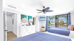 A bedroom with two beds, a ceiling fan, a TV, and a balcony with a view of palm trees and buildings. - Gertrude's Siesta | 6 Terraces, Rooftop, Pool, 1-min to Beach and Village! (Siesta Key)