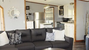 En moderne stue med en mørkegrå hjørnesofa, et sofabord og et køkkenområde, der ses gennem en åben dør.