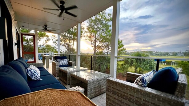 A covered outdoor seating area with wicker furniture and a glass table, overlooking a landscape with trees and a sunset. - Private Pool and Dock, Steps from the Beach, Beautiful Sunset Views (Saint Helena Island)