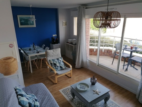 A living room with a sofa, a dining table with chairs, a coffee table with a decorative bowl and starfish, a blue wall, and a large window with a view of the outdoors.