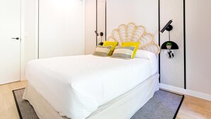 Een net opgemaakt bed met een witte dekbedovertrek en gele kussens, een wandlamp en een kleine potplant op een plank.
