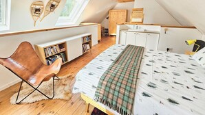 A cozy attic bedroom with a sloped ceiling, wooden beams, a bed with a plaid blanket, a bookshelf, a leather chair, and a small window.