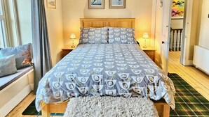Une chambre à coucher avec un grand lit, un lustre, une tête de lit en bois, deux tableaux encadrés au mur et une vue sur une autre pièce par une porte ouverte.