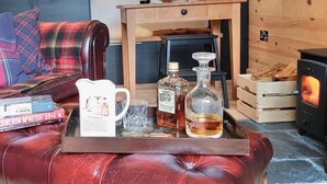A cozy living room with a leather sofa, a wooden coffee table with books and bottles, a fireplace, and a rustic wooden wall.