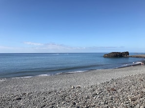 Uma praia de seixos com um afloramento rochoso ao longe e mar calmo.