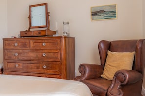 Quarto com penteadeira de madeira, poltrona de couro marrom com almofada e um quadro na parede.