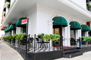 Un hotel con suelo a cuadros, mesas exteriores con toldos verdes y un cartel de 'For Sale'.