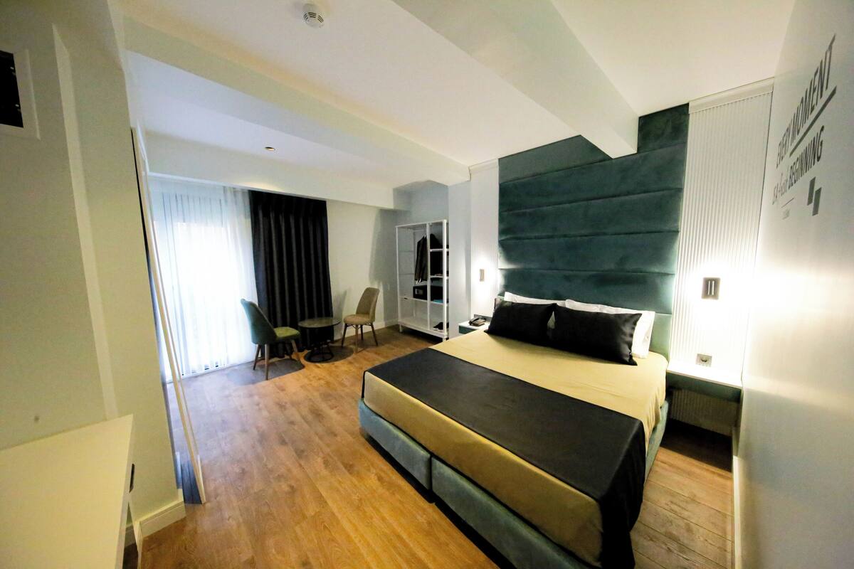 A modern hotel room with a large bed, a seating area with a chair and table, and a view of the outside through a window.