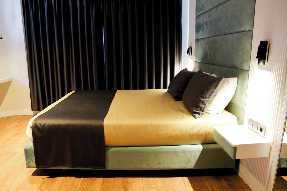 A modern bed with a green and beige bedspread, a white nightstand with a lamp, and a black curtain in the background.
