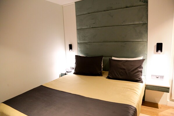 Una cama bien hecha con cabecera, dos mesitas de noche y una lámpara fijada a la pared.
