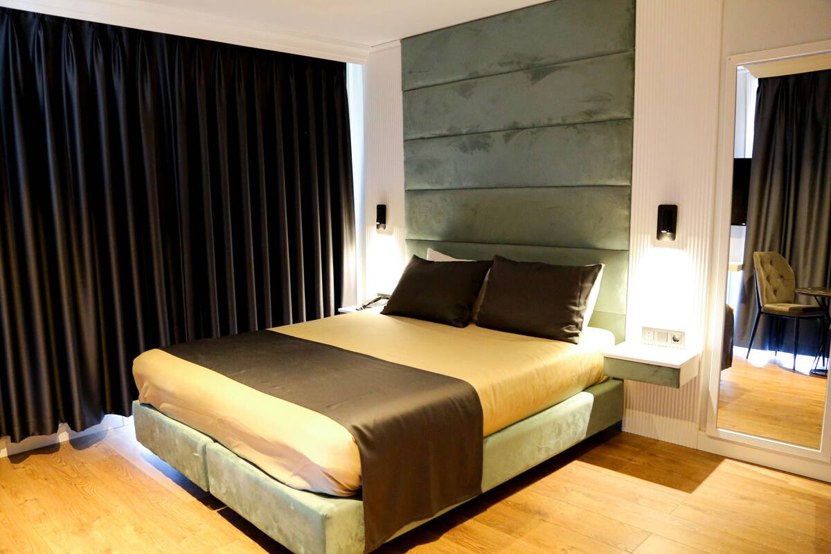 A modern hotel room with a large bed, a bedside table, a mirror, and a view of the room through a door.