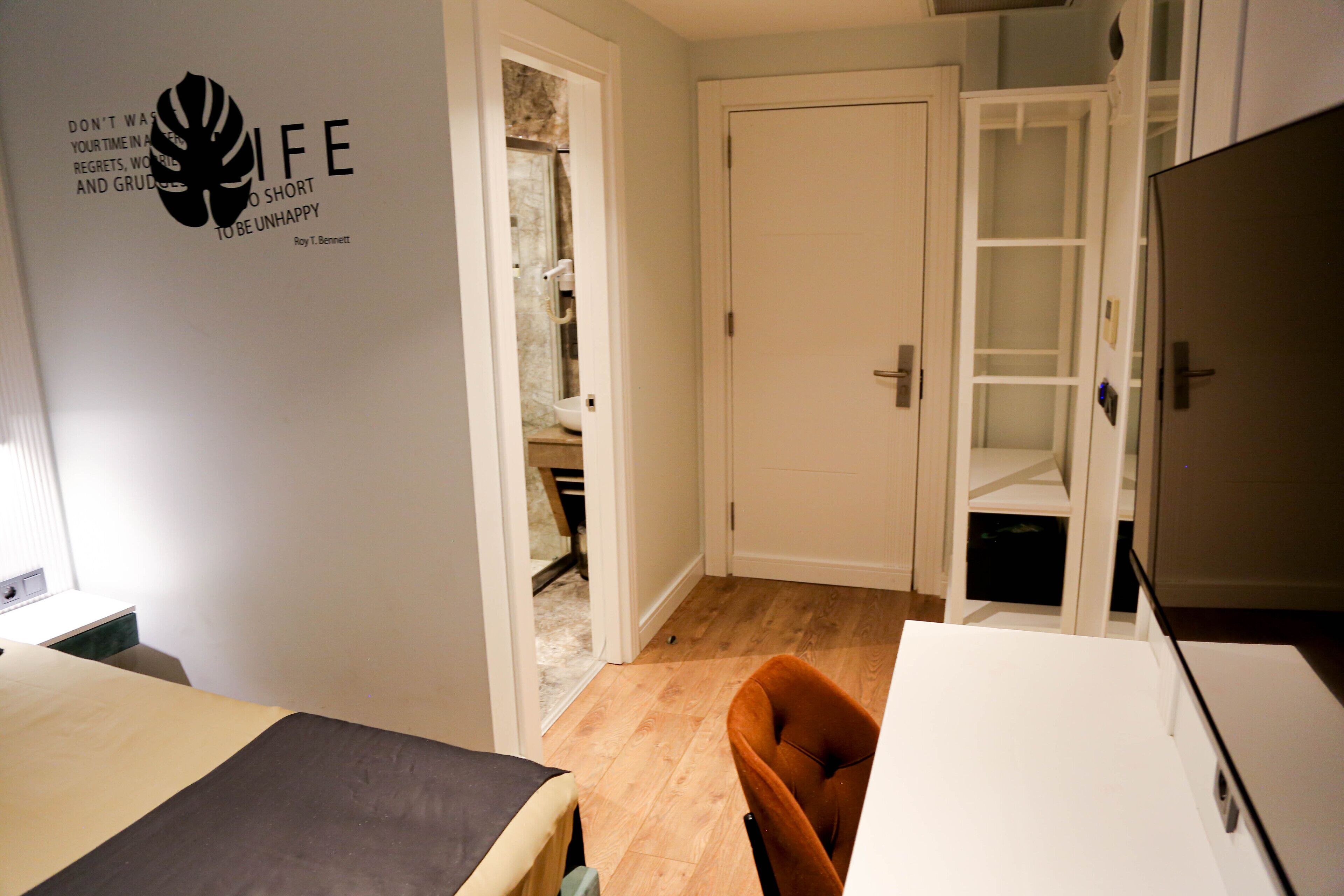 A hotel room with a bed, a chair, a desk, and a wardrobe. There is a door leading to a bathroom and a closet.