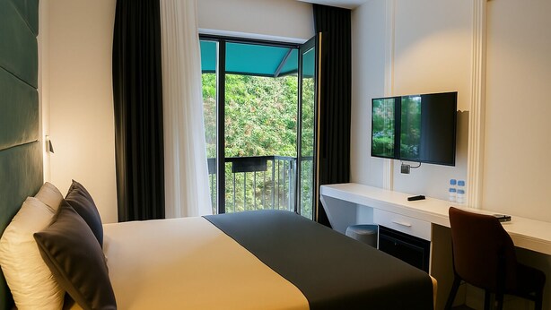 A hotel room with a bed, a desk, a chair, a TV, and a balcony with a view of greenery. - Axu Airport Hotel (Arnavutköy)