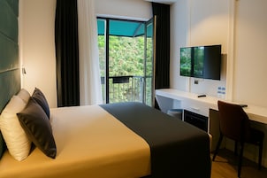 Habitación de hotel con cama, escritorio, silla, televisor y balcón con vista a follaje.