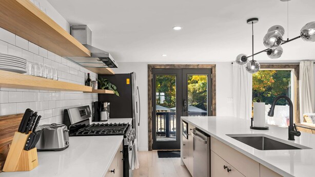 Eine moderne Küche mit weißen Arbeitsplatten, einem eingebauten Backofen und einer Spüle. Ein Fenster bietet Blick auf Bäume, und eine Tür führt in einen Außenbereich.