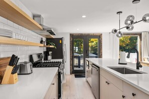 Una cocina moderna con encimeras blancas, horno integrado y un lavamanos. Se aprecia una ventana con vistas a árboles y una puerta que da a un espacio exterior.