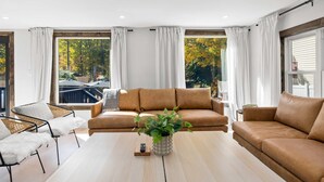 Ein modernes Wohnzimmer mit braunem Sofa, Couchtisch und Pflanze. Große Fenster mit Vorhängen bieten Blick auf Bäume und einen Balkon.