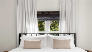Ein Bett mit weißer Bettwäsche und zwei beigen Kissen, ein schwarzes Metall-Kopfteil und weiße Vorhänge, die ein Fenster mit Blick auf Bäume umrahmen.