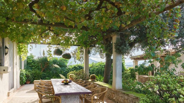 An outdoor patio with a wooden table and wicker chairs, surrounded by greenery and a stone wall.