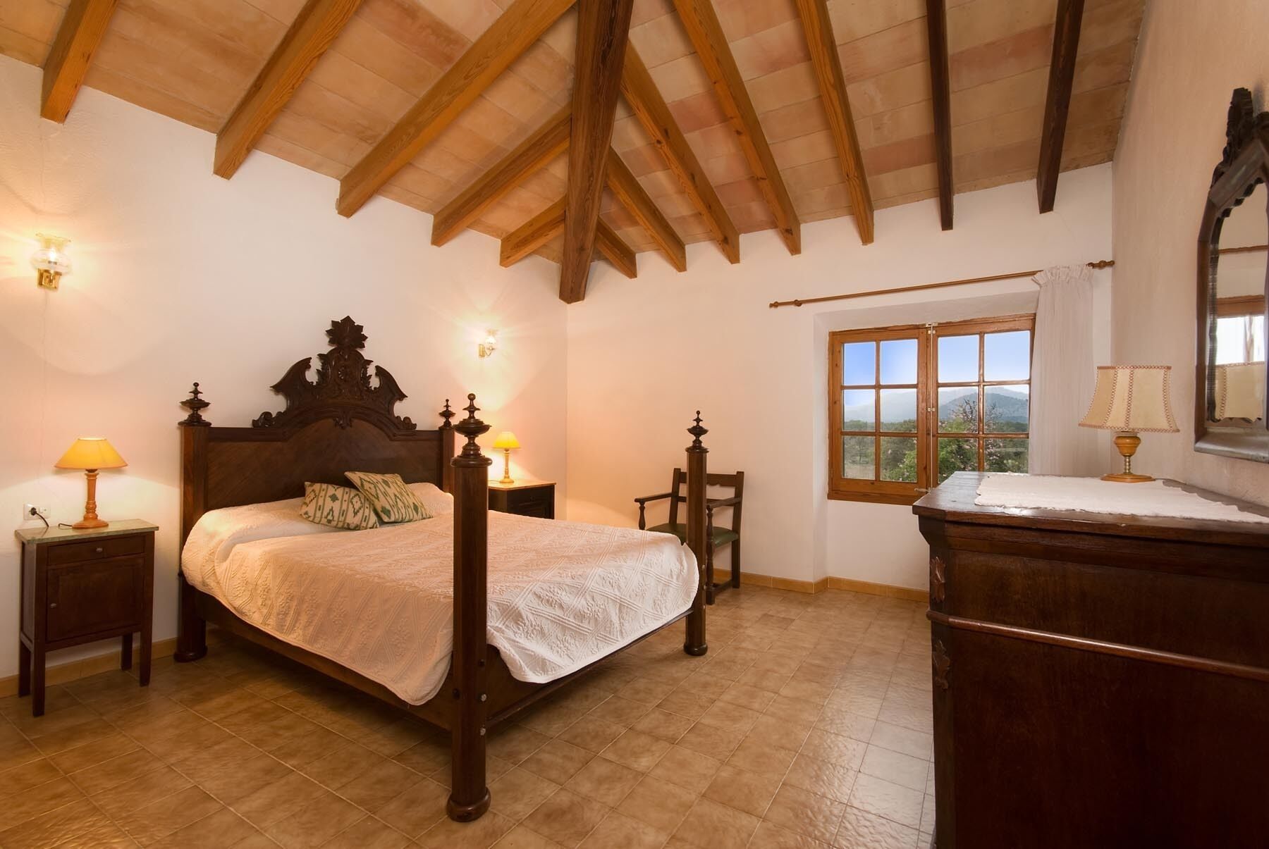 A bedroom with a wooden bed, two bedside tables with lamps, a chair, a dresser, and a window with a view of the mountains.