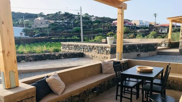 A covered outdoor seating area with stone walls, a table, and chairs. - The cave of benikulà ancient Pantelleria Dammuso Andrea 2 (Scauri)