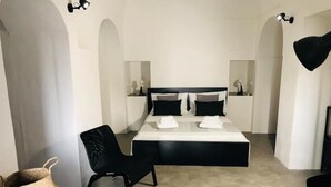 A modern hotel room with a large bed, a black chair, a small table, and a woven basket. - The cave of benikulà ancient Pantelleria Dammuso Andrea 2 (Scauri)