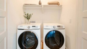 Two white Samsung front-loading washing machines in a laundry room with a small plant on the left and a shelf above. - Nashville music-themed house sleeps 10 near Belmont Mansion (Nashville)