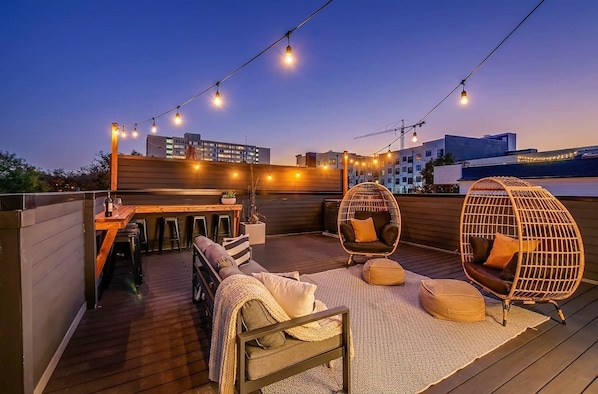 A rooftop terrace with a wooden deck, seating area with a sofa and chairs, string lights, and a view of buildings in the background.
