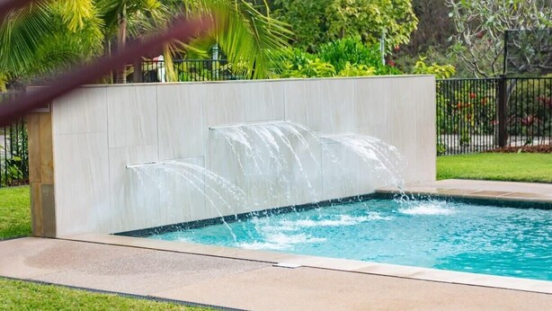 Una piscina con una cascada, rodeada por un muro de color claro y una zona cubierta de césped.