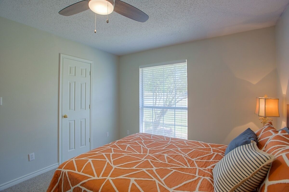Une chambre à coucher comprenant un lit, un ventilateur de plafond, une fenêtre avec des stores et une table de chevet avec une lampe.
