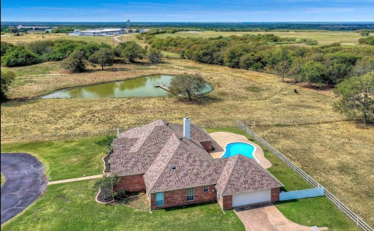 Une maison à deux étages avec une piscine, entourée d’une cour clôturée et d’un étang.