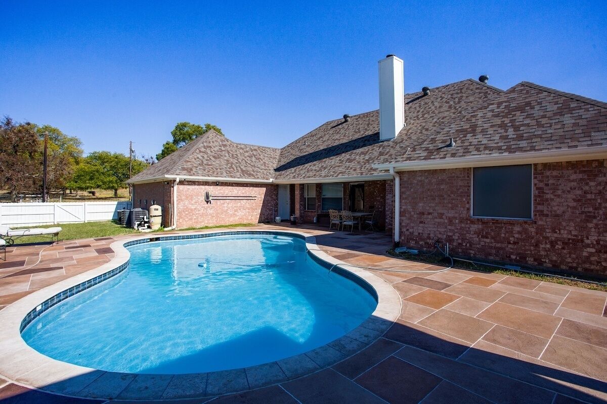 A backyard with a swimming pool, patio area, and a brick house with a chimney.