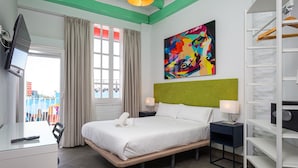 Ein Hotelzimmer mit Bett, Nachttischen, Fernseher, einem Fenster mit Vorhängen, einem farbenfrohen abstrakten Gemälde und Blick ins Freie.
