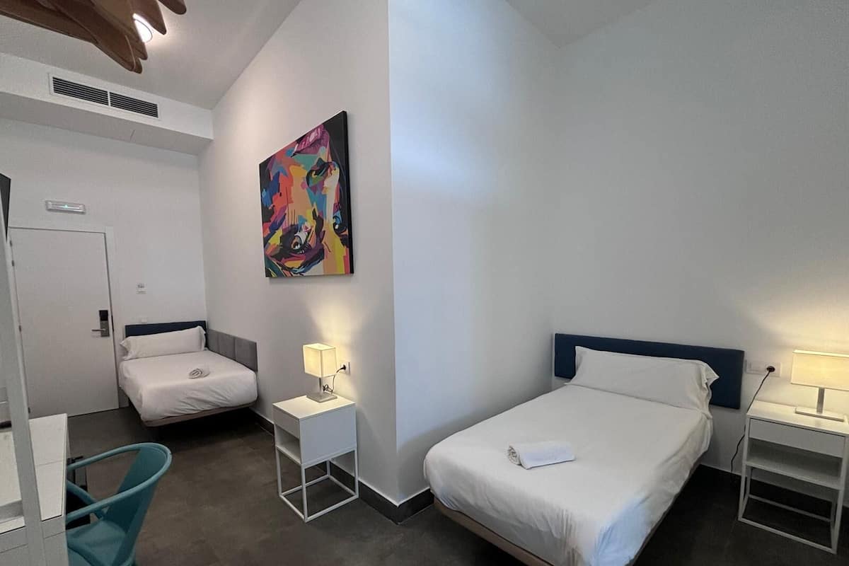 Une chambre d’hôtel avec deux lits simples, un bureau, une chaise, un tableau au mur et une porte.