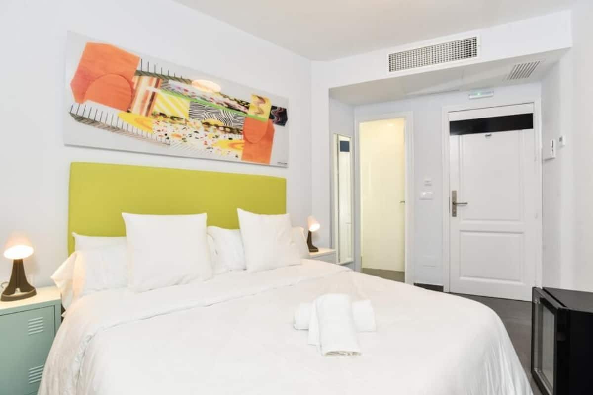 Une chambre d’hôtel avec un lit, du linge de lit blanc, une tête de lit verte, un tableau au mur, une porte et un téléviseur.