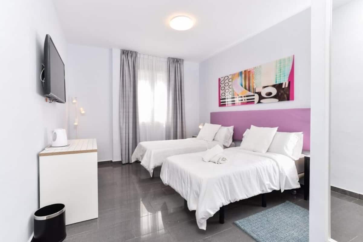 Une chambre d’hôtel avec deux lits, un téléviseur à écran plat, une machine à café, une poubelle et une peinture abstraite aux couleurs vives accrochée au mur.
