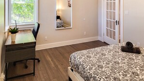 Un dormitorio con cama, escritorio con silla, planta en maceta, ventana con persianas, espejo y puerta.