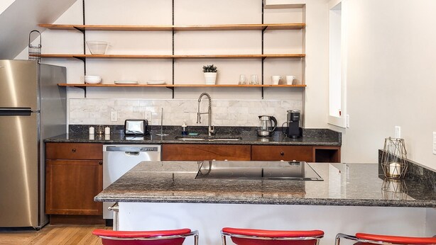 Una cocina moderna con estantes de madera, un refrigerador de acero inoxidable, lavavajillas integrado, encimera de granito, un lavamanos y tres taburetes rojos.