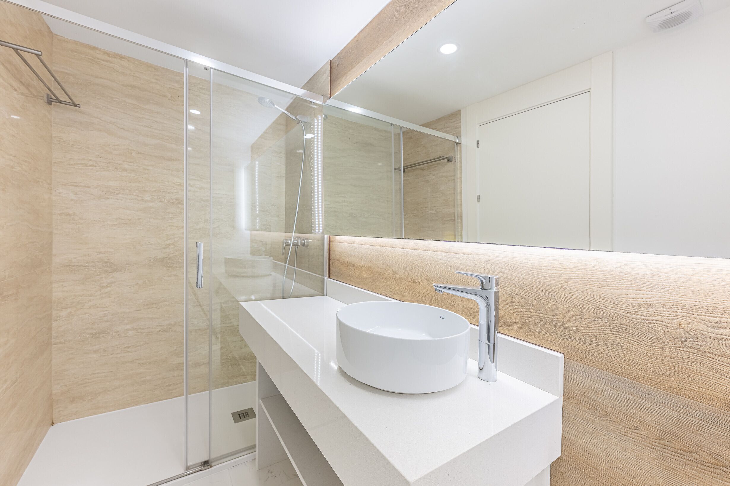 Une salle de bain moderne avec une douche à parois de verre, un lavabo blanc doté d’un plan de toilette en bois et un grand miroir.