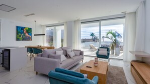 Een moderne woonkamer met een grijze bank, blauwe fauteuils, een houten salontafel, en een eethoek met een witte tafel en stoelen.