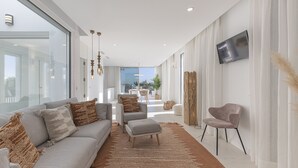 A modern living room with a grey sofa, a small grey armchair, and a matching ottoman. A flat-screen TV is mounted on the wall, and a large window provides natural light. - Villa EDEN 2399 private pool & sea view (San Roque)