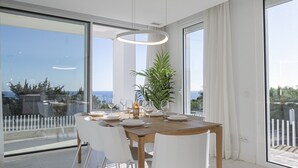 A modern dining area with a wooden table, white chairs, and a large window offering a view of the sea. - Villa EDEN 2399 private pool & sea view (San Roque)
